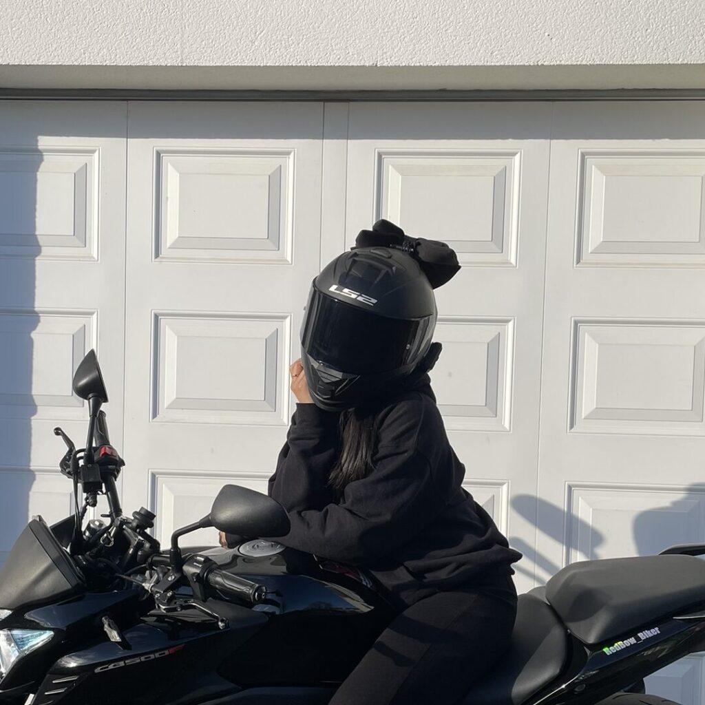 Motarde RedBow assise sur une moto noire avec un noeud de casque noir et un sweat à capuche noir sur un fond blanc